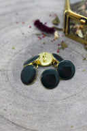 Cinq boutons ronds facettés vert forêt très foncé cerclés d'un liseré doré, disposés en cercle autour d'un anneau de couture doré visible, sur fond de pierre grise avec fleurs séchées.