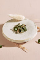 Boutons ronds bombés vert lierre cerclés d'un fin liseré doré, à queue métallique (shank), présentés en tas dans une petite coupelle en céramique blanche sur fond rose, avec un brin d'herbe séchée (lagurus) en arrière-plan.