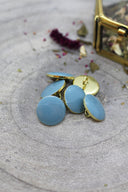 Five faceted round buttons in light blue-grey (storm) rimmed with a gold trim, with a visible gold sewing shank ring, on grey stone with dried flowers and a blurred gold box in the background.