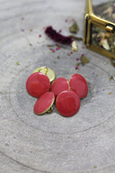 Close-up of five domed, faceted round buttons in glossy terracotta (orange-red), rimmed with a thin gold trim, set on textured grey stone with dried flowers and a small blurred gold box in the background.