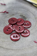 Fully glittered two-hole round buttons in burgundy, piled on grey stone, with a strand of burgundy raffia in the background.