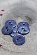 Fully glittered two-hole round buttons in midnight blue, piled on grey stone, with a strand of burgundy raffia in the background.