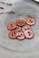 Boutons ronds à deux trous entièrement pailletés cuivre rosé, présentés en tas sur une pierre grise, avec un fil de raphia bordeaux en arrière-plan.