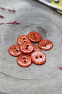 Boutons ronds à deux trous entièrement pailletés orange rouille, présentés en tas sur une pierre grise, avec un fil de raphia bordeaux en arrière-plan.