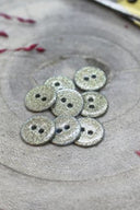 Boutons ronds à deux trous entièrement pailletés doré pâle champagne, présentés en tas sur une pierre grise, avec un fil de raphia bordeaux en arrière-plan.