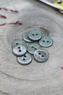 Boutons ronds à deux trous entièrement pailletés gris argenté, présentés en tas sur une pierre grise, avec un fil de raphia bordeaux en arrière-plan.