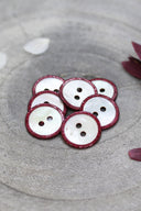 Two-hole round buttons in white mother-of-pearl, rimmed with a glittery burgundy amarante trim, piled in a grey stone dish, with white and burgundy flower petals in the background.