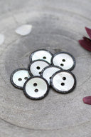 Two-hole round buttons in white mother-of-pearl, rimmed with a glittery black trim, piled in a grey stone dish, with white and burgundy flower petals in the background.