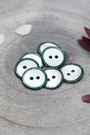 Two-hole round buttons in white mother-of-pearl, rimmed with a glittery cactus green trim, piled in a grey stone dish, with white and burgundy flower petals in the background.
