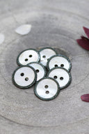 Two-hole round buttons in white mother-of-pearl, rimmed with a glittery dark green trim, piled in a grey stone dish, with white and burgundy flower petals in the background.