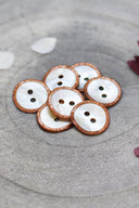 Two-hole round buttons in white mother-of-pearl, rimmed with a glittery copper chestnut trim, piled in a grey stone dish, with white and burgundy flower petals in the background.