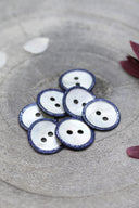 Two-hole round buttons in white mother-of-pearl, rimmed with a glittery cobalt blue trim, piled in a grey stone dish, with white and burgundy flower petals in the background.