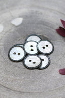 Two-hole round buttons in white mother-of-pearl, rimmed with a glittery anthracite trim, piled in a grey stone dish, with white and burgundy flower petals in the background.