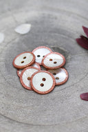 Two-hole round buttons in white mother-of-pearl, rimmed with a glittery rose copper trim, piled in a grey stone dish, with white and burgundy flower petals in the background.