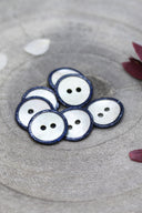 Two-hole round buttons in white mother-of-pearl, rimmed with a glittery navy blue trim, piled in a grey stone dish, with white and burgundy flower petals in the background.