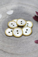 Two-hole round buttons in white mother-of-pearl, rimmed with a glittery mustard gold trim, piled in a grey stone dish, with white and burgundy flower petals in the background.