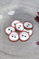 Boutons ronds à deux trous en nacre blanche, cerclés d'un liseré pailleté rouge terracotta, présentés en tas dans une coupelle en pierre grise, avec des pétales de fleurs blanches et bordeaux en arrière-plan.