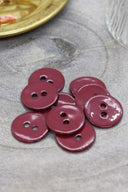 Two-hole round buttons in glossy burgundy amarante enamel, piled on grey stone, with a gold dish of dried flowers and a faceted glass in the background.
