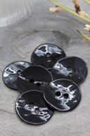 Boutons ronds à deux trous en émail brillant noir, présentés en tas sur une pierre grise, avec une coupelle dorée contenant des fleurs séchées et un verre facetté en arrière-plan.