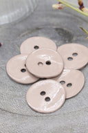 Boutons ronds à deux trous en émail brillant rose blush, présentés en tas sur une pierre grise, avec une coupelle dorée contenant des fleurs séchées et un verre facetté en arrière-plan.