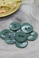 Boutons ronds à deux trous en émail brillant vert cactus, présentés en tas sur une pierre grise, avec une coupelle dorée contenant des fleurs séchées et un verre facetté en arrière-plan.
