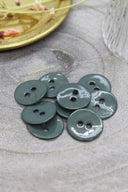 Two-hole round buttons in glossy cedar green enamel, piled on grey stone, with a gold dish of dried flowers and a faceted glass in the background.