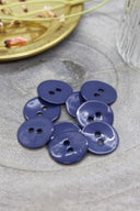 Two-hole round buttons in glossy cobalt blue enamel, piled on grey stone, with a gold dish holding dried flowers and a faceted glass in the background.