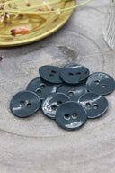 Two-hole round buttons in glossy dark forest blue-grey enamel, piled on grey stone, with a gold dish holding dried flowers and a faceted glass in the background.