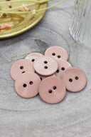Boutons ronds à deux trous en émail brillant rose beige sable, présentés en tas sur une pierre grise, avec une coupelle dorée contenant des fleurs séchées et un verre facetté en arrière-plan.