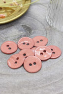 Two-hole round buttons in glossy melba pink enamel, piled on grey stone, with a gold dish holding dried flowers and a faceted glass in the background.