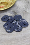 Two-hole round buttons in glossy navy blue enamel, piled on grey stone, with a gold dish holding dried flowers and a faceted glass in the background.