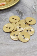 Two-hole round buttons in glossy mustard enamel, piled on grey stone, with a gold dish holding dried flowers and a faceted glass in the background.