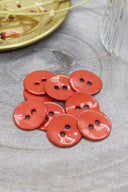 Boutons ronds à deux trous en émail brillant orange corail vif, présentés en tas sur une pierre grise, avec une coupelle dorée contenant des fleurs séchées et un verre facetté en arrière-plan.