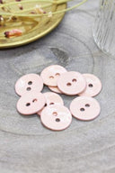 Two-hole round buttons in glossy very pale powder pink enamel, piled on grey stone, with a gold dish holding dried flowers and a faceted glass in the background.