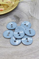 Boutons ronds à deux trous en émail brillant bleu gris pâle storm, présentés en tas sur une pierre grise, avec une coupelle dorée contenant des fleurs séchées et un verre facetté en arrière-plan.