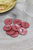 Boutons ronds à deux trous en émail brillant rouge terracotta, présentés en tas sur une pierre grise, avec une coupelle dorée contenant des fleurs séchées et un verre facetté en arrière-plan.