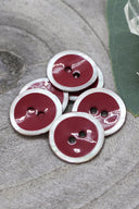 Boutons ronds à deux trous, finition émaillée rouge amarante cerclée d'un liseré blanc nacré, présentés en tas sur une pierre grise, avec une feuille verte et une petite coupelle en verre facetté en arrière-plan.