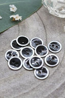 Two-hole round buttons, black enamel finish rimmed with a pearly white trim, piled on grey stone, with a green leaf and a small faceted glass dish in the background.