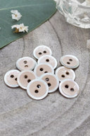 Boutons ronds à deux trous, finition émaillée rose blush cerclée d'un liseré blanc nacré, présentés en tas sur une feuille verte, avec une petite coupelle en verre facetté et des pétales de fleurs blanches en arrière-plan.