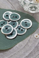 Boutons ronds à deux trous, finition émaillée vert cactus cerclée d'un liseré blanc nacré, présentés en tas sur une feuille verte, avec une petite coupelle en verre facetté et des pétales de fleurs blanches en arrière-plan.
