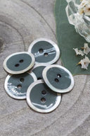 Two-hole round buttons, cedar grey enamel finish rimmed with a pearly white trim, piled on grey stone, with a green leaf and a small faceted glass dish in the background.