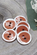 Two-hole round buttons, auburn chestnut brown enamel finish rimmed with a pearly white trim, piled on grey stone, with a green leaf and a small faceted glass dish in the background.
