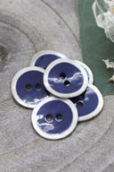 Two-hole round buttons, cobalt blue enamel finish rimmed with a pearly white trim, piled on grey stone, with a green leaf and a small faceted glass dish in the background.