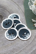 Two-hole round buttons, dark forest green enamel finish rimmed with a pearly white trim, piled on grey stone, with a green leaf and a small faceted glass dish in the background.