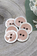 Boutons ronds à deux trous, finition émaillée beige rosé sable cerclée d'un liseré blanc nacré, présentés en tas sur une feuille verte, avec une petite coupelle en verre facetté et des pétales de fleurs blanches en arrière-plan.