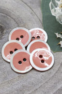 Two-hole round buttons, melba pink enamel finish rimmed with a pearly white trim, piled on grey stone, with a green leaf and a small faceted glass dish in the background.