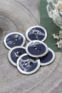 Two-hole round buttons, midnight blue enamel finish rimmed with a pearly white trim, piled on grey stone, with a green leaf and a small faceted glass dish in the background.