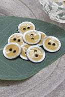 Two-hole round buttons, mustard enamel finish rimmed with a pearly white trim, piled on a green leaf, with a small faceted glass dish and white flower petals in the background.