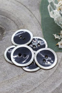 Two-hole round buttons, anthracite grey enamel finish rimmed with a pearly white trim, piled on grey stone, with a green leaf and a small faceted glass dish in the background.