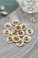 Two-hole round buttons, ochre enamel finish rimmed with a pearly white trim, piled on a green leaf, with a small faceted glass dish and white flower petals in the background.